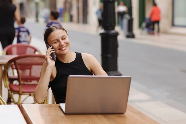 Successful smiling young business woman communication at smartphone during works on laptop at cafe outdoors. Concept of remote work from public place, digital freelance and modern lifestyle.