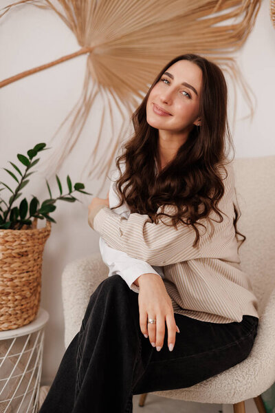 Attractive confident young stylish brunette woman with long hair looks at camera at living room with creative design in rustic style. Beauty, fashion, self-expression, individuality concept