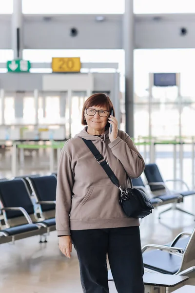 Happy smiling mature traveler woman hold smartphone and wave hand in greeting someone who meeting her at airport terminal after arrived from trip at new country at holiday vacation. Retirement travel.
