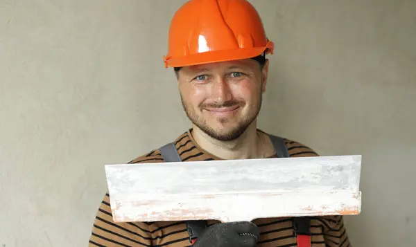 Smiling handyman repairman worker in overalls and protective orange helmet holds big putty knife for plastering concrete wall with putty. Renovation at apartment and construction new building concept
