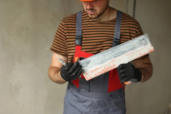 Worker in overalls and safety hard hat holds and prepares two spatulas for plastering concrete wall with putty using. Renovation apartment and construction new building concept. Copy place.