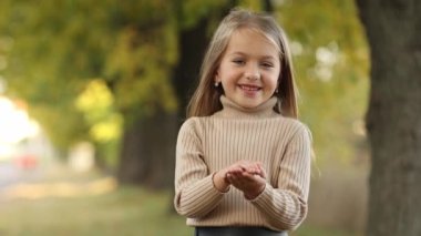Çocuklar Günü 'nde. Neşeli küçük kız 5-6 yaşında güler ve sonbahar parkında yapraklarla oynar. Kafkasyalı anaokulu öğrencisi çocuk örgü süveter giyiyor. Sonbahar tatilinde boş zaman aktivitesi