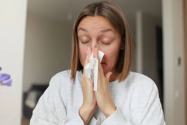 Seasonal allergy. Close up face of allergic young woman uses paper tissues from runny nose, sneezes from flowers pollen at home. Girl with close eyes with flu, itching or cough, allergy rhinitis.