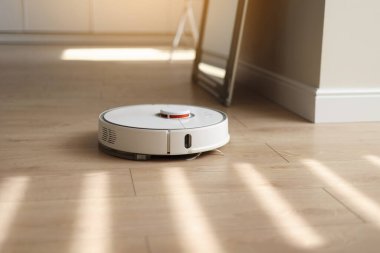 Yuvarlak kablosuz otonom robot elektrikli süpürge. Ahşap zemin üzerinde güneş ışığı var. Ev kumandası aletleri. Akıllı temizlik teknolojisi. Otomatik itişli temizlik robotu. Yer temizleme sistemi.