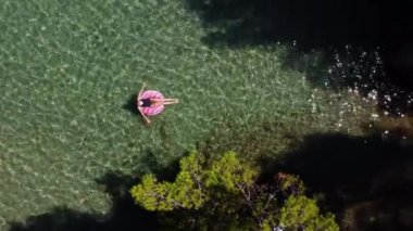 Açık kollu, mayo giymiş, yüzen ve dinlenen genç bir kadının havadan drone görüntüsü. Su yüzeyinde şeffaf bir tabanla yüzüyor. Egzotik tatil beldesinde yaz tatili.