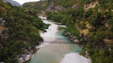 Karadağ 'ın turkuaz nehri ile güzel kayalık bir kanyonun ortasındaki asma eski metal köprünün hava aracı görüntüsü. Moraca nehri kanyonu yeşil canlı bitki örtüsüyle