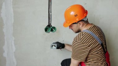 Üniformalı elektrikçi erkek, koruyucu eldivenler ve kask prizdeki elektrik voltajını kontrol ediyor elektrikli tornavida kullanıyor. Kapalı alandaki voltaj detektörüyle prizdeki kabloları inceliyor..