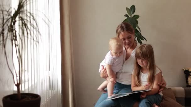 ママは自宅でソファーにいる小さな娘たちのために本を読んだ 幼稚園の少女は 彼女の妹と母親との時間と魅力的なおとぎ話の休息を楽しんでいます キッズ開発 知的成長コンセプト — ストック動画