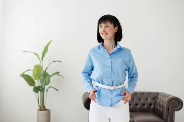 Women equality day. Smiling successful businesswoman in elegant blue shirt and white pants looks at camera at modern comfortable light office with green plant and sofa. Work life balance concept