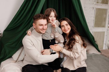 Mutlu aile günleri. Genç, sevgi dolu ebeveynler, anne-baba ve şirin bir anaokulu çocuğu olan küçük kız çocuğu, eğleniyor, rahat bir yatak odasında kameraya bakarak kucaklaşıyor. Ebeveynlik çocukluk kavramı