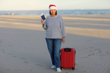 Noel Baba şapkalı gülümseyen olgun kadın sahilde pasaport, bilet ve kırmızı bavul taşıyor. Mutlu yıllar 2024 bayram kutlamaları. Kış tatili