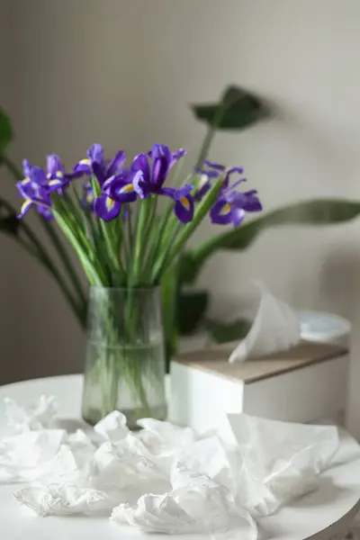 Concept of seasonal allergy to flower. Bouquet of purple iris flowers in a glass vase on table and many used napkins at home interior. Allergic rhinitis symptom of odor pollen flowering.