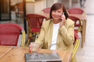 Olgun bir ofis çalışanı, elinde bir fincan kahve ve dizüstü bilgisayarla terasta otururken akıllı telefonuyla yaklaşan iş seyahatinden bahseder.