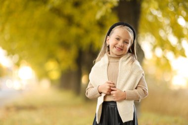 Çocuk günü. 5-6 yaşlarında neşeli bir kız çocuğu. Sonbahar parkındaki ağacın yanında durup kameraya poz veriyor ve gülümsüyor. Şefkatli kreş çocuğu bej rengi örgü süveter giyiyor. Mutlu çocukluk