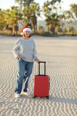 Noel Baba şapkalı, çizgili tişörtlü, 50-55 yaş arası mutlu bir yetişkin kadın kum sahili kenarında kırmızı bavul tutuyor. Palmiyeli yeni yıl tatilini tropikal tatil beldesinde yalnız kutluyor..
