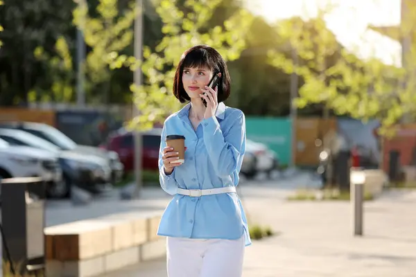 Work life balance. Adult confident elegant businesswoman talks on smartphone, holds cup of take away coffee outdoors on city street. Business concept, urban people, city lifestyle working people