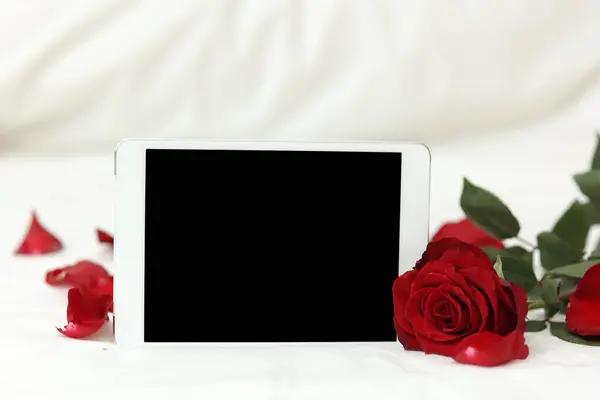 Tablet with black screen mockup for modern electronic invitation or greeting card, red rose flower and petals on white background. St. Valentines day celebration. Women's day. Copy space for text