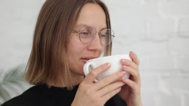 Kahve molası. 20-30 yıl boyunca sıcak bir şeyler içen genç bir kadın. Beyaz gözlüklü beyaz bir kız elinde beyaz bir fincan kahve, kafeteryada çay, sosyal medyayı okuyor, gülümsüyor..