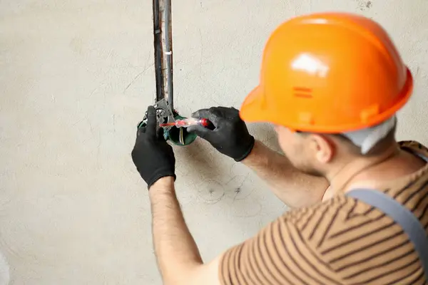 Electrician male in uniform, protective gloves and helmet checks presence of electrical voltage in socket phase uses electrical tester screwdriver.Examining wires in outlet by voltage detector indoor.