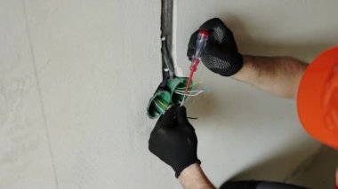 Üniformalı elektrikçi erkek, koruyucu eldivenler ve kask prizdeki elektrik voltajını kontrol ediyor elektrikli tornavida kullanıyor. Kapalı alandaki voltaj detektörüyle prizdeki kabloları inceliyor..