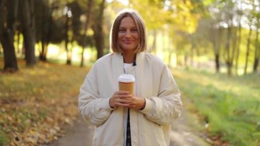 Gülümseyen şık, kısa saçlı genç bir kadın elinde eko-kağıt bardakta sıcak içecek tutuyor, kahve ya da çay, kameraya bakıyor, renkli sonbahar parkında soğuk bir günün tadını çıkarıyor. Mutlu sıfır israf yaşam tarzı konsepti.