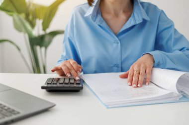 Unrecognisable businesswoman calculates on calculator expenses, fees, analyses financial reports, does accounting, paperwork job, laptop on desk at office. Investment, savings, currency concept. clipart