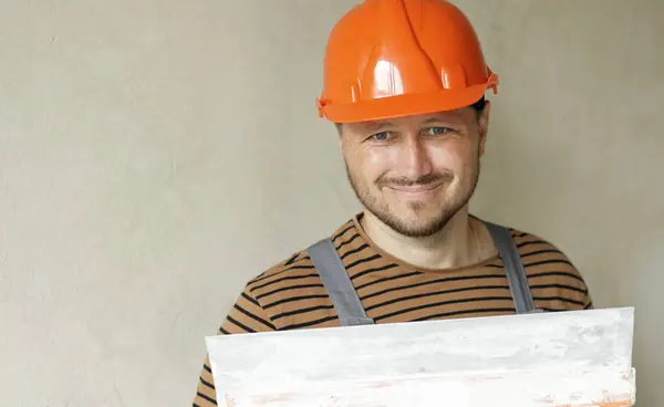 Smiling handyman repairman worker in overalls and protective orange helmet holds big putty knife for plastering concrete wall with putty. Renovation at apartment and construction new building concept