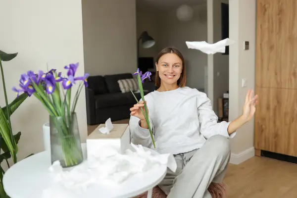 stock image Allergy free. Cheerful young woman throws napkins up at home. Happy life without of runny nose, itching or cough symptoms. Allergen bouquet of iris flowers at vase, used paper handkerchiefs on table.