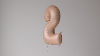 Yumuşak bej hasırın 2 numaralı balon şekli açık arkaplanda hareket ediyor. Bebek partisi için helyum doğum günü balonu dönüyor ve uçuyor. Modern kutlama yıldönümü tatili.