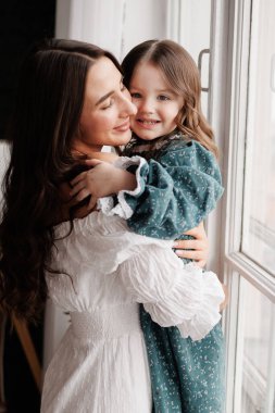 Anneler Günü. Mutlu genç anne, sevimli kızıyla pencerenin önünde kucaklaşıyor. Anneyle çocuk arasındaki güven, destek ve sevgi. Sevgi dolu aile tatlı anların tadını çıkarır. Çocuk günü