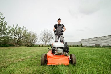 Koruyucu giyimli profesyonel bahçıvan arka bahçedeki modern benzin kablosuz çim biçme makinesini kullanarak yeşil çimen biçiyor. Mevsimlik peyzaj tasarımı. Arkaplanda çiçek açan ağaçlar.