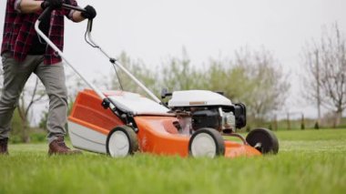 Tanımlanamayan bir erkek, gündelik giysiler içinde modern benzin kablosuz çim biçme makinesini itiyor, evinin arka bahçesindeki yeşil çimenleri buduyor ve umursuyor. Bahçe bakımı aletleri ve ekipmanları.