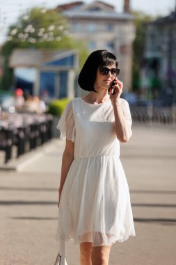 Kısa saçlı romantik esmer kadın mini beyaz elbise ve güneş gözlüğü takıyor, cep telefonuyla konuşuyor, yazın kalabalık bir caddede yürüyor. Kız dışarıda akıllı telefonla iletişim kuruyor..
