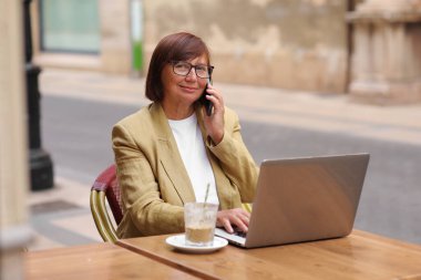 Olgun bir ofis çalışanı, elinde bir fincan kahve ve dizüstü bilgisayarla terasta otururken akıllı telefonuyla yaklaşan iş seyahatinden bahseder.