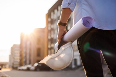 Unrecognisable engineer, architect or worker holds in hand protective white hard hat and blueprint on multi-apartment residential complex background, sunlighting. Concept of new real estate clipart