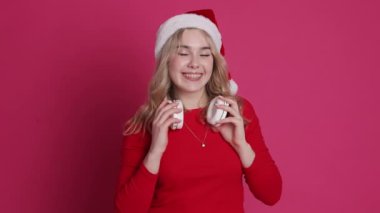 Neşeli genç sarışın kadın Noel Baba şapkası takıyor ve kablosuz kulaklık takıyor, pembe arka plan üzerinden izole edilmiş kameraya gülümsüyor ve flört ediyor. Mutlu yıllar 2025 ve Xmas kış tatillerini kutluyor..
