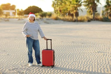 Noel Baba şapkalı, çizgili tişörtlü, 50-55 yaş arası mutlu bir yetişkin kadın kum sahili kenarında kırmızı bavul tutuyor. Palmiyeli yeni yıl tatilini tropikal tatil beldesinde yalnız kutluyor..