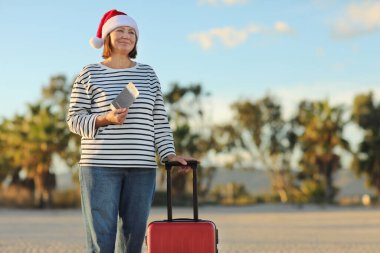Noel Baba şapkalı, çizgili tişörtlü, bağımsız olgun bir kadın pasaport, bilet, el fonları ve deniz kenarında çanta taşıyor. Yeni yılı egzotik tatil beldelerinde kutluyor..