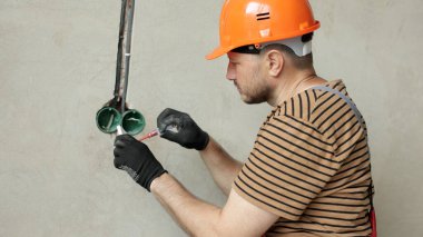 Üniformalı elektrikçi erkek, koruyucu eldivenler ve kask prizdeki elektrik voltajını kontrol ediyor elektrikli tornavida kullanıyor. Kapalı alandaki voltaj detektörüyle prizdeki kabloları inceliyor..