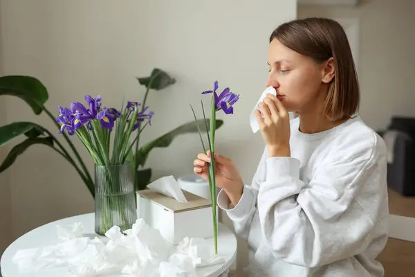 stock image Allergic young woman holds iris flower, covers nose with paper tissue has runny nose, sneezes from flowers pollen at kitchen home by table. Girl with flu, itching or cough, seasonal allergy, rhinitis.