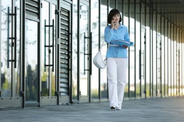 Çok görevli genç iş kadını, yürüyüş sırasında akıllı telefonla iletişim kuruyor. Önemli finans belgelerini dışarıdaki ofis binalarına bırakıyor. Stres direnci ve iş konsepti..