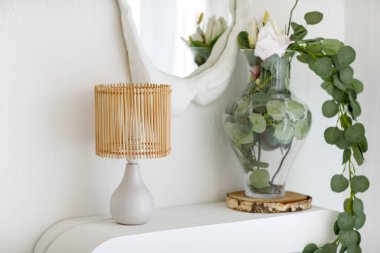 İskandinav minimalist iç tasarımı masa rattan lambasıyla, okaliptüs dalları cam vazoda ve ayna ile aydınlık oturma odasındaki duvarda dokulu beyaz alçı çerçeve. Konforlu ev dekorasyonu