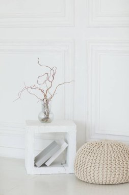 Küf çerçeveli beyaz duvarlı, aydınlık oturma odasının iç tasarımı bej işlemeli pouf, beton küp masa üzerindeki cam vazodaki doğal ağaç dalları, kitaplar. İskandinav modern ev dekorasyonu.