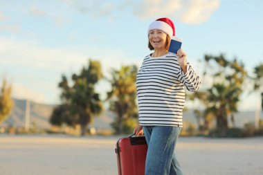 Noel Baba şapkalı neşeli yetişkin kadın kırmızı bavul, pasaport ve bilet taşıyor. Sahilde palmiyelerle yürüyor. Yeni yılı egzotik tatil beldelerinde kutluyor.