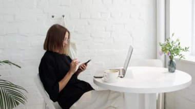 Serbest çalışan genç bir kadın akıllı telefon kullanıyor, internette geziniyor, sohbet ediyor, gülümsüyor, şık bir kafede dizüstü bilgisayarla oturuyor. Modern teknoloji, gençlik yaşam tarzı. Uzaktan çalışma, e-öğrenme veya çevrimiçi uzaktan çalışma.