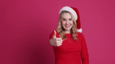 Noel Baba şapkalı dişlek sarışın kadınla gülümseyen genç, pembe arka planda izole edilmiş bir jest gibi başparmağını kaldırıyor. Mutlu yıllar 2025 ve Xmas kış tatillerini kutluyor.