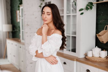 Kadınlar Günü. 20-25 yaşlarında, uzun kıvırcık saçlı, çapraz elli romantik ve çekici bir genç kadın, arka plandaki modern moda ev mutfağına bakıyor.