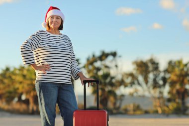 Noel Baba şapkalı, çizgili tişörtlü, 50 'li yaşlarda neşeli olgun bir kadın plajda kırmızı bavul tutuyor ve 2024 kış tatilini tropik tatil beldesinde kutluyor. Boşluğu kopyala.
