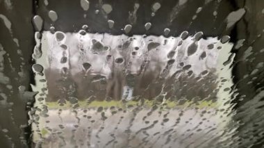 İçerideki su akışı arabanın ön camındaki köpüğü ve kiri silip süpürüyor. Otomatik oto yıkama işlemi. Araba camında sabun köpükleri. Beyaz sabun köpüğü araba camında hareket eder..