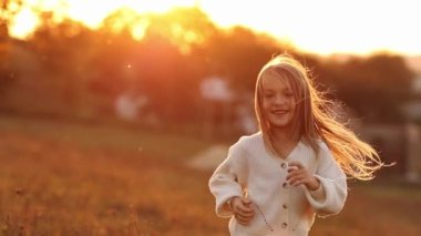 Sevimli neşeli kız 5-6 yıl boyunca gülümseyerek koştu ve gün batımında dışarıda kameraya baktı. Gündelik giysiler içindeki çocuk, sonbahar tatilinde doğada dinlenmenin keyfini çıkarır. Mutlu çocukluk kavramı
